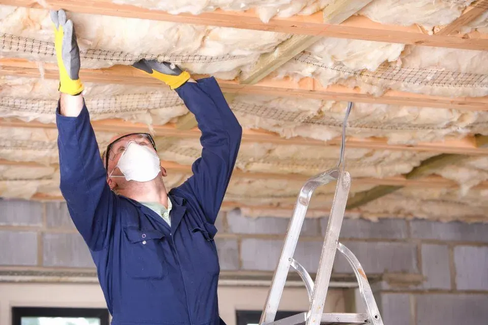 Installer fitting glasswool ceiling batts between joists from a ladder